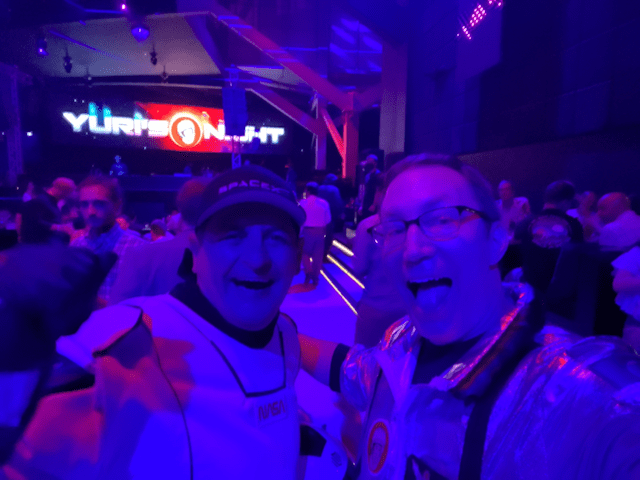 Mac Malkawi and Tim Bailey smile in flight suits in a large, dark dance club. There is purple lighting barely illuminating the scene with a giant Yuri's Night sign in the back.