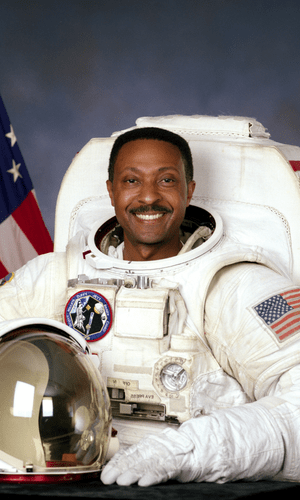 Retired NASA Astronaut Winston Scott, a black man with short black hair and a black mustache, smiling in his bulky, white Extra Vehicular Activity suit